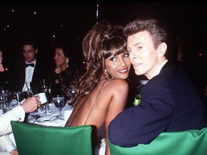 David Bowie z Iman siedzą przy stole, odwracając się i patrząc w kamerę